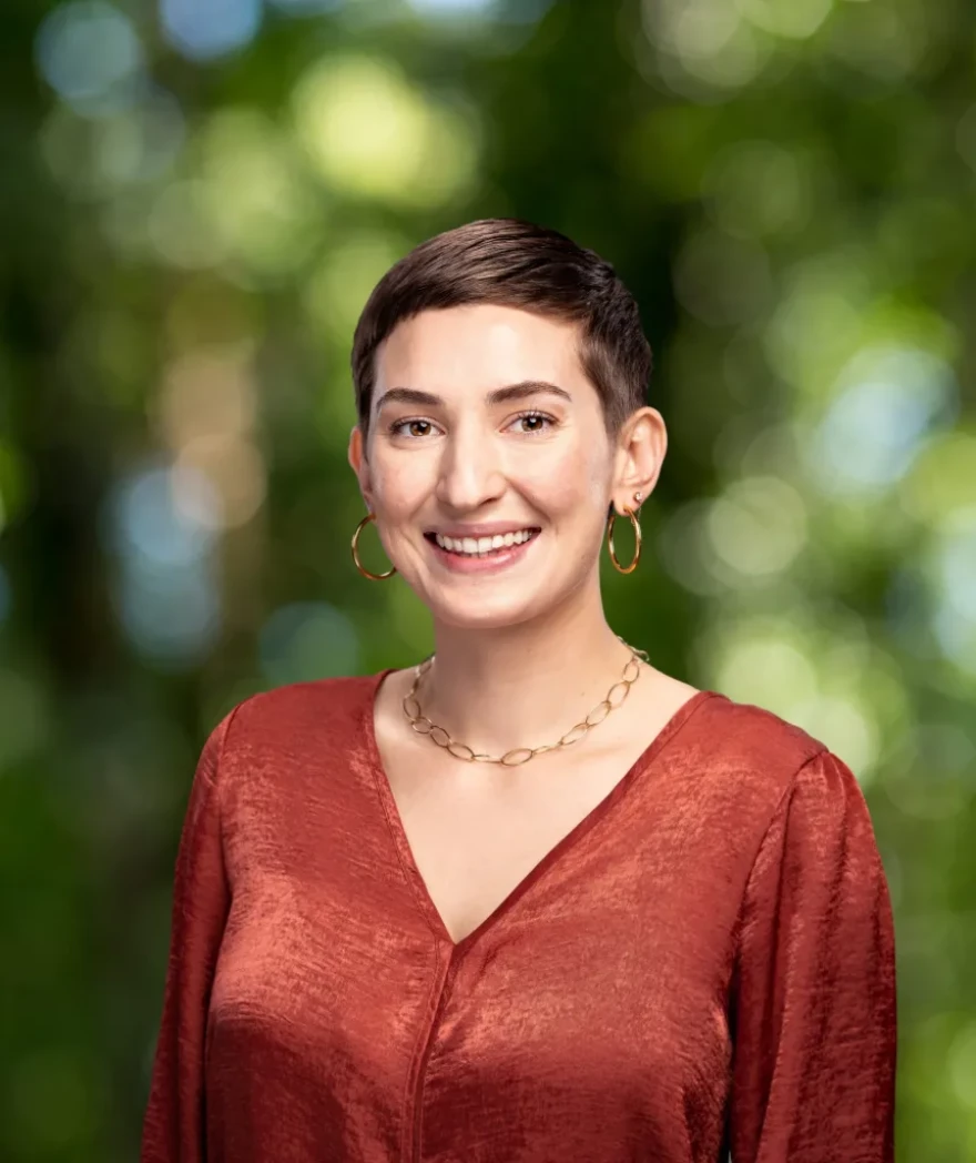 A white woman, Erin Walton-Haggerty, pictured here with short brown hair in a red shirt standing in front of a blurred green scenery.